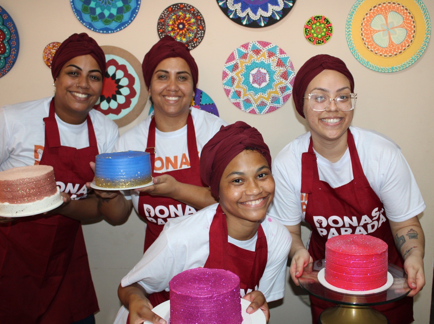 Quatro mulheres com aventais vermelhos segurando bolos e sorrindo